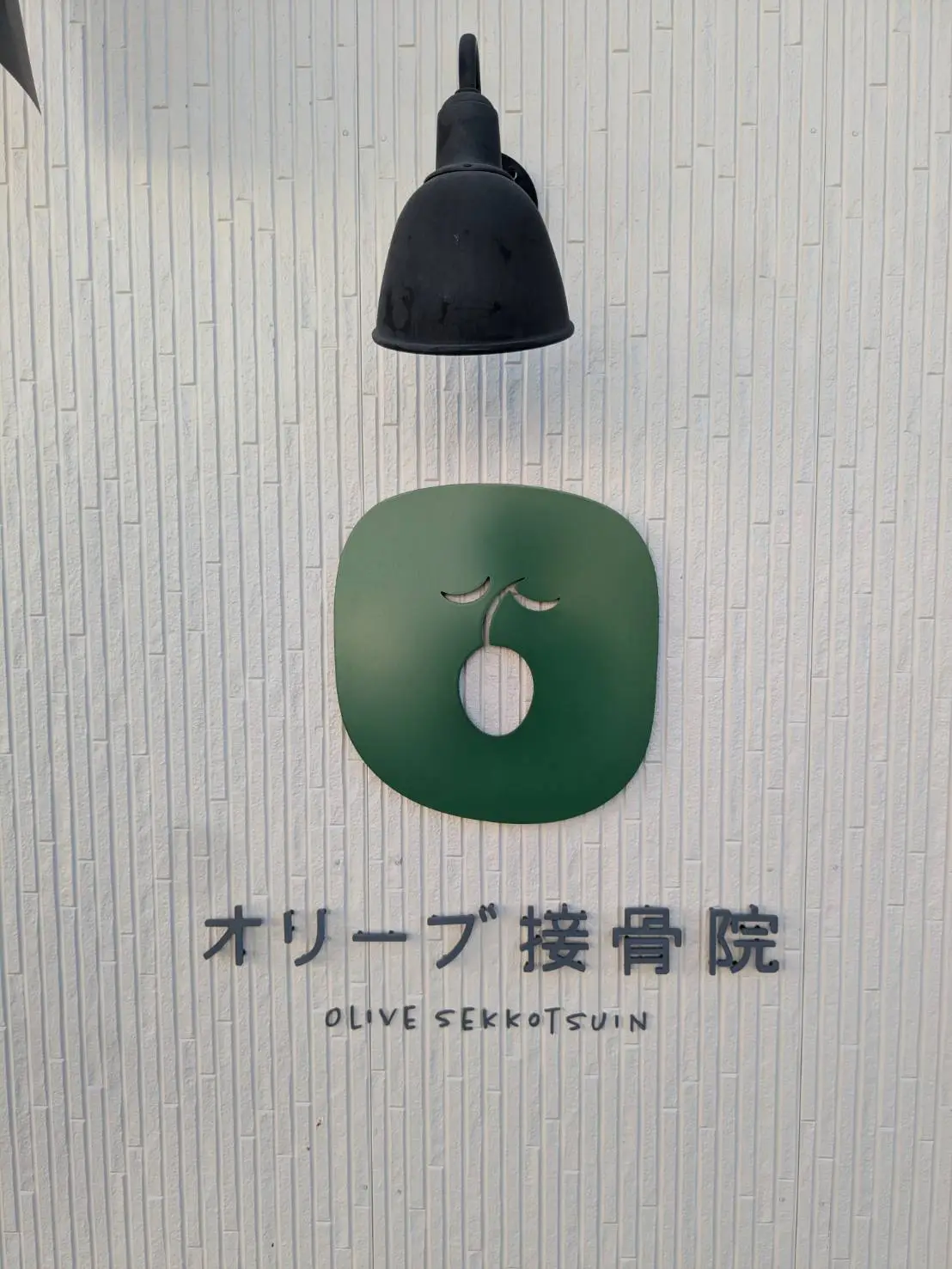 入口の壁に取り付けられた、オリーブのロゴマークと「オリーブ接骨院 OLIVE SEKKOTSUIN」のサイン。
